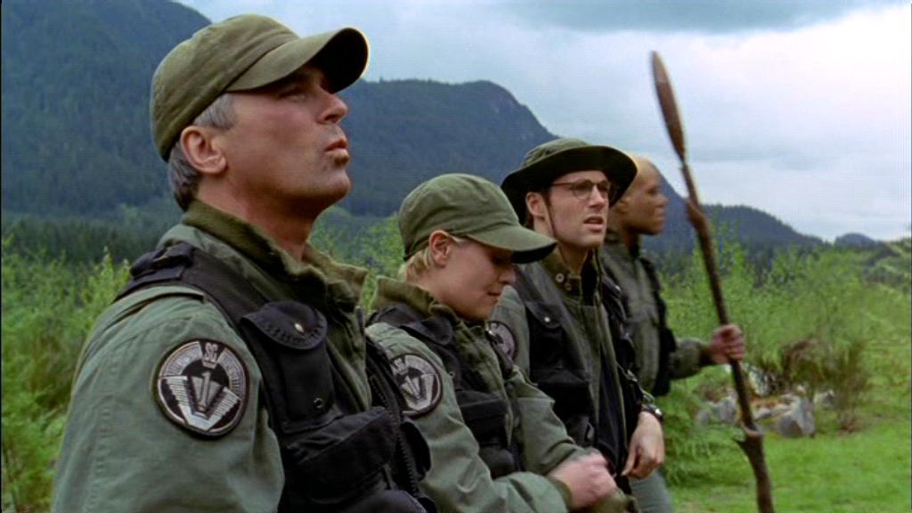 Jack O'Neill, Samantha Carter, Daniel Jackson, and Teal'c stand on one side of the stargate surrounded by trees and bushes in the Stargate SG-1 episode 'Demons'. Behind them a large moon is clearly visible in the sky, although it is daytime.