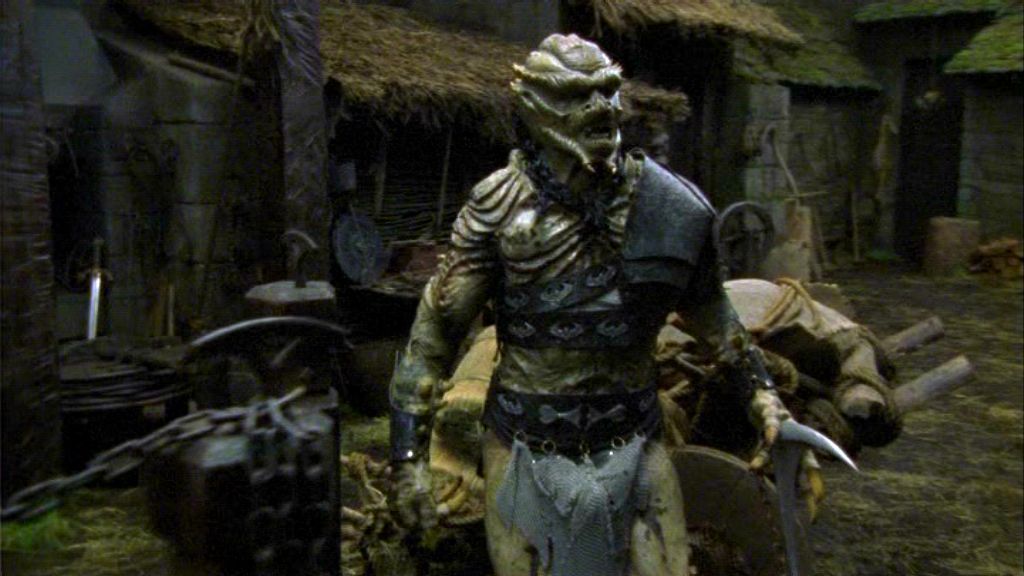 An alien Unas stands in a Medieval courtyard, a cart visible behind him and the floor earthen, in the Stargate SG-1 episode 'Demons'.