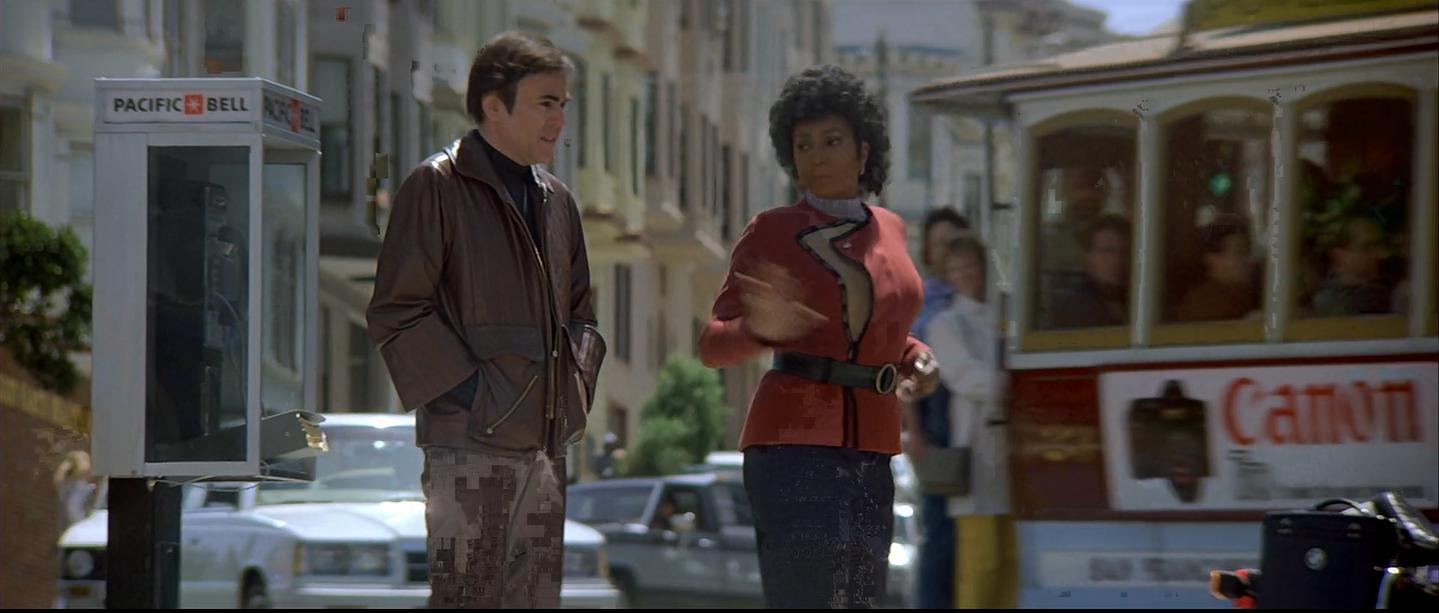 Uhura wearing a red tunic turning to Chekov. They are standing in the street with a San Francisco tram visible in the background of the scene.