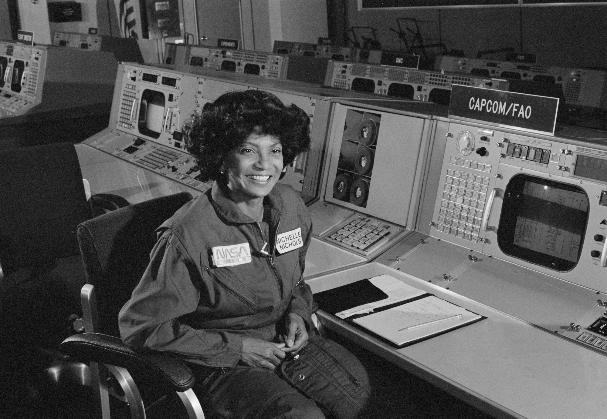 Nichelle Nichols sits in NASA mission control and smiles. She is wearing a flightsuit with her name on it.